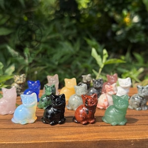 May include: A selection of small, carved cat figurines in diverse colours and materials, including pink, blue, green, black, and red. The cats are displayed on a wooden surface with a blurred green backdrop.