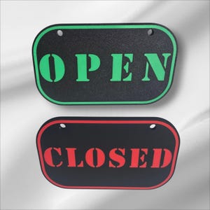 Peut inclure: Deux panneaux rectangulaires aux coins arrondis. Le panneau supérieur est noir avec un bord vert et le mot "OPEN" en vert. Le panneau inférieur est noir avec un bord rouge et le mot "CLOSED" en rouge. Chaque panneau a deux trous pour accrocher.