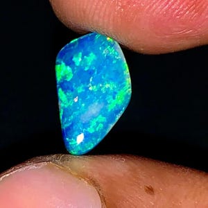 以下が含まれることがあります： 鮮やかな青色のオパール宝石で、緑と白の輝きがあります。磨かれた石は、2本の指の間に黒い背景を背景に保持され、独特の色合いを際立たせています。この宝石は、天然オパールの美しい例です。