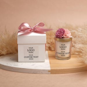 May include: A white gift box tied with a pink ribbon and a small candle topped with flowers. The box and candle both have space for a logo and custom text.