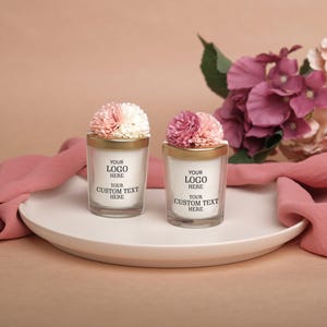 May include: Two glass candles with gold rims and floral decorations are displayed on a white plate. Each candle has text that reads "YOUR LOGO HERE" and "YOUR CUSTOM TEXT HERE". Pink fabric and flowers are in the background.