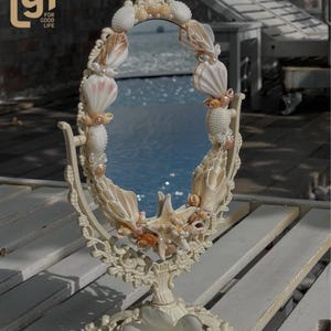 May include: An oval vanity mirror with a cream-colored frame, adorned with seashells, pearls, and a starfish. The mirror is set on a decorative stand with a floral base. The image includes the text "lgf FOR GOOD LIFE".