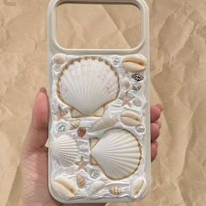 Può includere: Una cover per telefono color crema con una superficie bianca testurizzata decorata con varie conchiglie, piccole stelle marine e pietre levigate. Le conchiglie più grandi sono delineate da un sottile bordo color oro, creando un design a rilievo.
