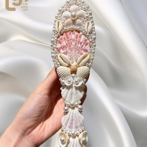 Op de afbeelding: Een ovaalvormig decoratief object gemaakt van schelpen en parels. Het ontwerp heeft een centrale roze en witte schelp, omringd door kleinere schelpen en parels. Het object wordt tegen een witte satijnen achtergrond gehouden. De tekst "lgf FOR GOOD LIFE" staat linksboven.