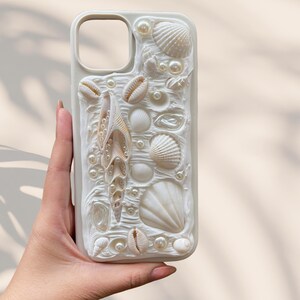 May include: A white phone case with a raised seashell design, including various shell shapes and faux pearls. The case has a cutout for the camera and the text "for good life" in the upper left corner.