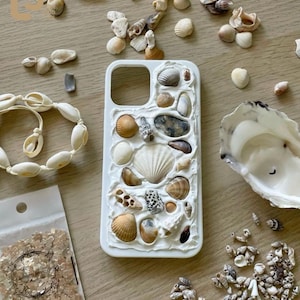 Może przedstawiać: Białe etui na telefon ozdobione różnymi muszlami, w tym muszlami i innymi stworzeniami morskimi. Obecna jest również bransoletka z muszli i małe torebki z elementami dekoracyjnymi, a także luźne muszle rozrzucone na drewnianej powierzchni.