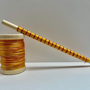 May include: A wooden spool of orange and yellow thread next to a wooden dowel wrapped with alternating red and yellow thread. The thread and dowel are for crafting and sewing projects.