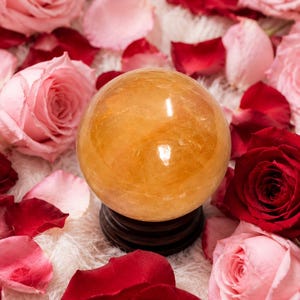May include: A polished, translucent golden-orange sphere made of natural crystal, resting on a dark brown circular wooden stand. The sphere is surrounded by a scattering of vibrant red and soft pink rose petals and several full rose blooms.