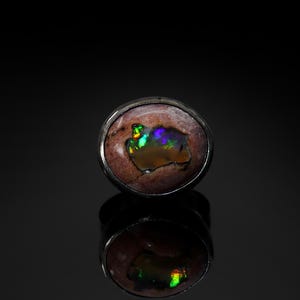May include: A round, silver-toned ring featuring a natural opal gemstone. The opal has a brown base color with iridescent flashes of green, blue, and yellow. The ring is set against a dark background, with its reflection visible below.