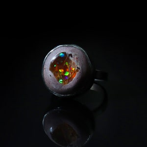 Può includere: Un anello d'argento con un grande opale ovale. L'opale mostra un vibrante gioco di colori, con lampi di arancione, verde e blu. L'incastonatura dell'anello è di colore viola chiaro e il gambo è in argento.