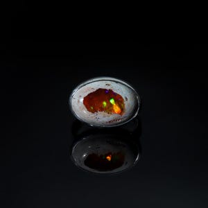 May include: Oval-shaped opal ring with a silver band. The opal features a vibrant display of colors, including red, orange, green, and purple, set against a white and brown background. The ring is displayed on a reflective black surface.