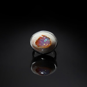 May include: A silver ring featuring a large, oval opal gemstone. The opal displays a mix of colors, including orange, purple, and green, set against a white background. The ring is set on a reflective black surface.
