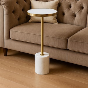 Puede incluir: Una mesa auxiliar moderna con tapa blanca y borde dorado. La mesa tiene un pie dorado y una base de mármol blanco. Se coloca junto a un sofá marrón con botones y un cojín beige.