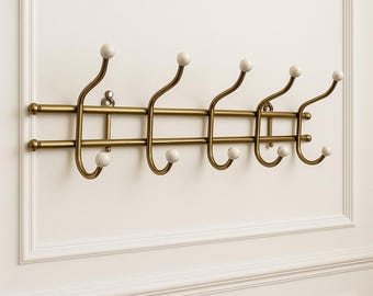 May include: A brass-colored wall-mounted coat rack with six hooks. Each hook has a curved design with a white, spherical knob at the end. The rack is mounted on a white wall with decorative molding.