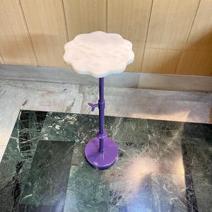 May include: A decorative, adjustable height side table. The table features a white, scalloped marble top and a purple metal base and stem. The base is round, and the stem has an adjustment mechanism. The table is designed for display.