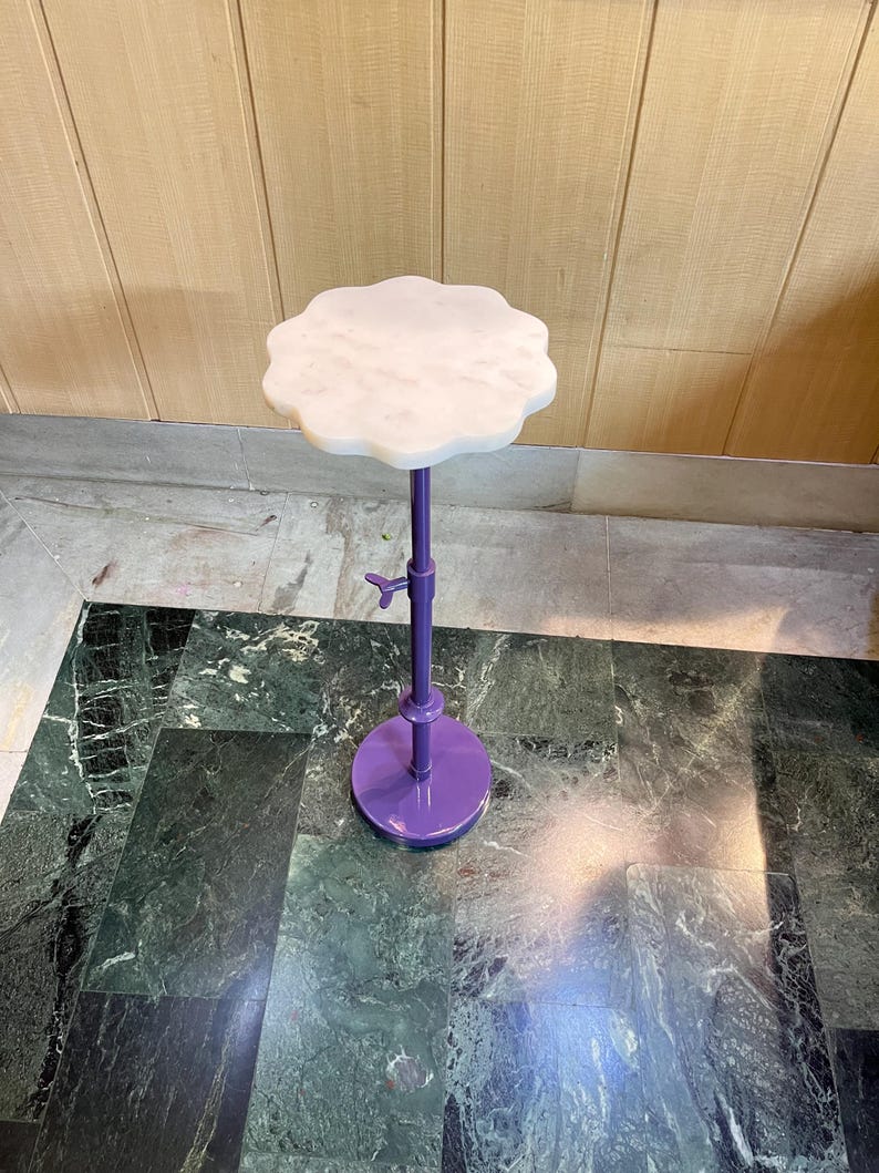 May include: A decorative, adjustable height side table. The table features a white, scalloped marble top and a purple metal base and stem. The base is round, and the stem has an adjustment mechanism. The table is designed for display.