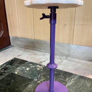 May include: A purple adjustable display stand with a round base and a cloud-shaped top. The stand has a central pole with a knob for height adjustment. The base and pole are a glossy purple, and the top is a light, off-white color.