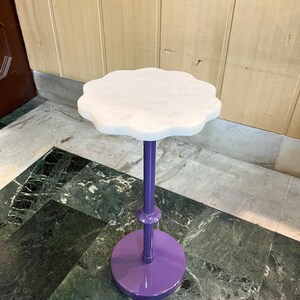 May include: A small, decorative plant stand with a white, scalloped marble top. The stand has a vibrant purple stem and base. The base is round, and the stem is a straight, slender pole. The stand is designed to hold a small plant or decorative item.