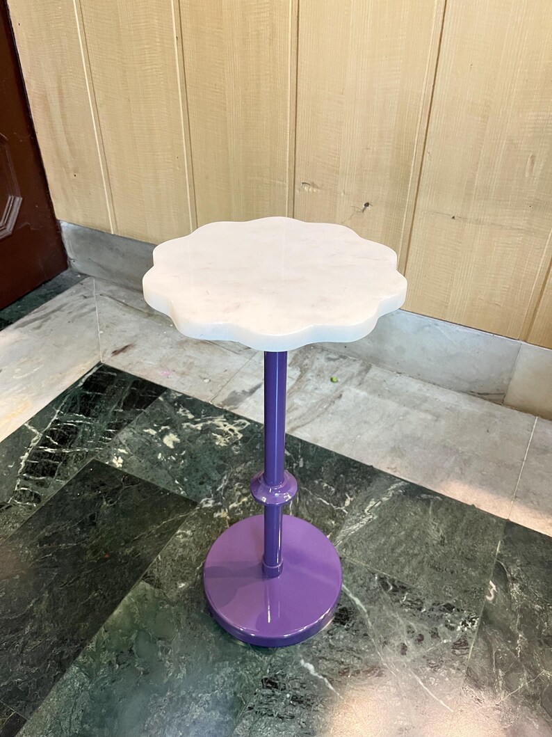 May include: A small, decorative plant stand with a white, scalloped marble top. The stand has a vibrant purple stem and base. The base is round, and the stem is a straight, slender pole. The stand is designed to hold a small plant or decorative item.