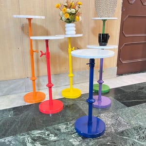 May include: A collection of colorful, adjustable side tables. Each table features a white circular top, a brightly colored stem, and a matching round base. Colors include orange, red, yellow, blue, purple, and green. Some tables hold decorative items.