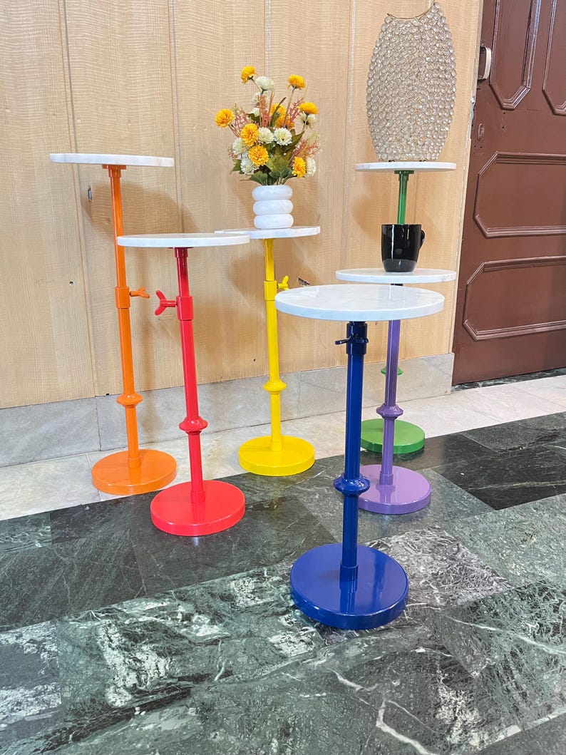 May include: A collection of colorful, adjustable side tables. Each table features a white circular top, a brightly colored stem, and a matching round base. Colors include orange, red, yellow, blue, purple, and green. Some tables hold decorative items.