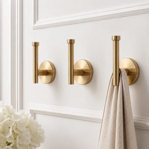 May include: Three gold-colored wall hooks with a circular base and a vertical bar. One hook holds a beige towel. The hooks are mounted on a white wall with decorative molding. A bouquet of white flowers is in the lower left corner.