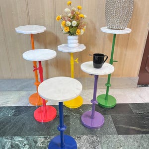 May include: A collection of colorful, small accent tables with white marble tops. The tables have bright bases in red, orange, yellow, purple, green, and blue. Some tables hold a vase, flowers, and a mug.