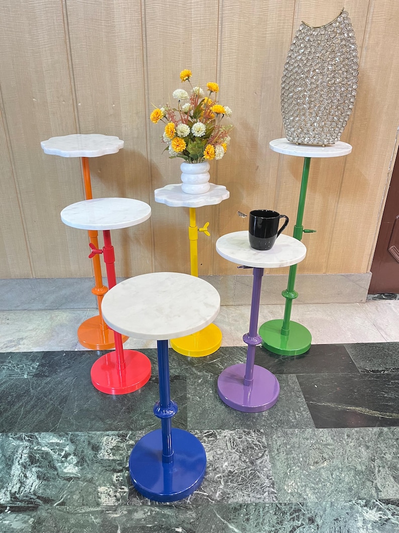May include: A collection of colorful, small accent tables with white marble tops. The tables have bright bases in red, orange, yellow, purple, green, and blue. Some tables hold a vase, flowers, and a mug.