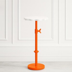 May include: A modern, adjustable side table with a white, scalloped top. The table has a bright orange stem and base, and a hand-turn adjustment knob. The round base is 12 inches in diameter.