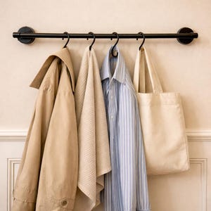 Könnte beinhalten: Eine schwarze Metall-Wandgarderobe mit einem beigefarbenen Trenchcoat, einem beigen Waffelhandtuch, einem blau-weiß gestreiften Hemd und einer cremefarbenen Tragetasche. Schlichtes, minimalistisches Design.