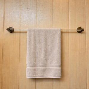 May include: A beige towel hangs from a clear acrylic and bronze towel rail. The rail is mounted on a light wood-panelled wall. The towel has a textured appearance with a decorative border.