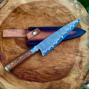 May include: A Damascus steel chef's knife with a wooden handle and a leather sheath. The blade has a wavy edge and a blue and silver pattern. The handle is brown and silver. The knife is resting on a wooden surface.