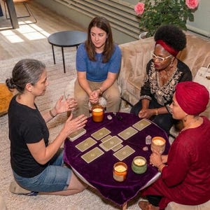 May include: Four individuals sit around a round table covered with a purple velvet cloth, featuring a spread of rectangular tarot cards, three lit candles in glass jars, and small crystals arranged in a cross-like pattern on the surface.
