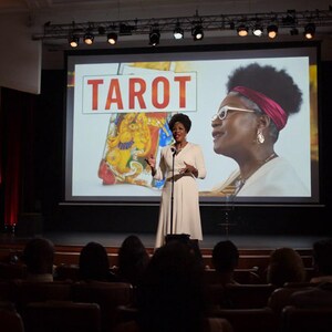 Op de afbeelding: Een persoon staat op een podium voor een groot projectiescherm met het woord TAROT in rode letters, een kleurrijke ge&iuml;llustreerde kaart en een grote afbeelding van een persoon met een wit montuur bril en een rode hoofdband.