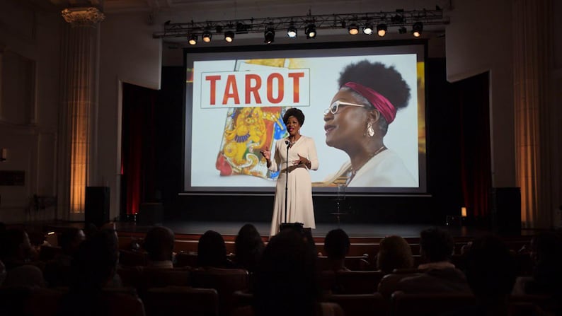 Op de afbeelding: Een persoon staat op een podium voor een groot projectiescherm met het woord TAROT in rode letters, een kleurrijke ge&iuml;llustreerde kaart en een grote afbeelding van een persoon met een wit montuur bril en een rode hoofdband.