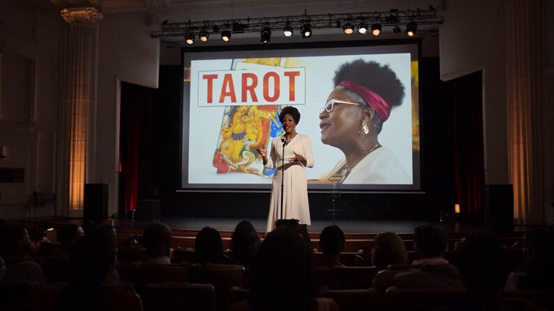 May include: A person stands on a stage in front of a large projection screen displaying the word TAROT in red letters, a colorful illustrated card, and a large image of a person wearing white-rimmed glasses and a red headband.
