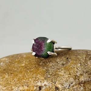May include: A silver ring featuring a raw, uncut gemstone with a mix of deep red and green hues. The stone is held in place by four prongs, and the ring is set on a light-colored stone.