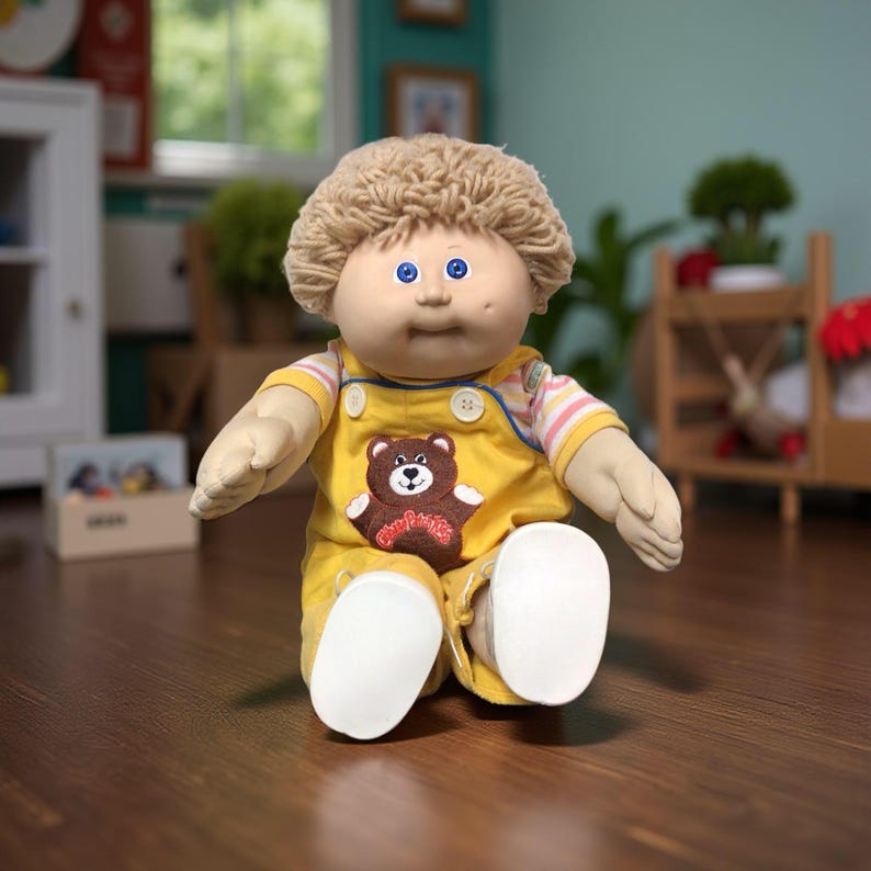 K&ouml;nnte beinhalten: Eine Vintage-Cabbage-Patch-Kids-Puppe mit weichem K&ouml;rper. Die Puppe hat ein rundes Gesicht, blaue Augen und hellbraunes, lockiges Haar. Sie tr&auml;gt einen gelben Overall mit einem braunen B&auml;renaufn&auml;her und wei&szlig;en Schuhen. Die Puppe sitzt auf einer Holzoberfl&auml;che.