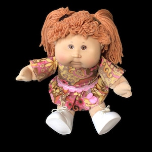 Puede incluir: Mu&ntilde;eca vintage con cabello de hilo naranja peinado en coletas. La mu&ntilde;eca viste un vestido con estampado floral en tonos rosas y marrones, y zapatos blancos. La mu&ntilde;eca tiene una cara redonda y est&aacute; sobre un fondo negro.