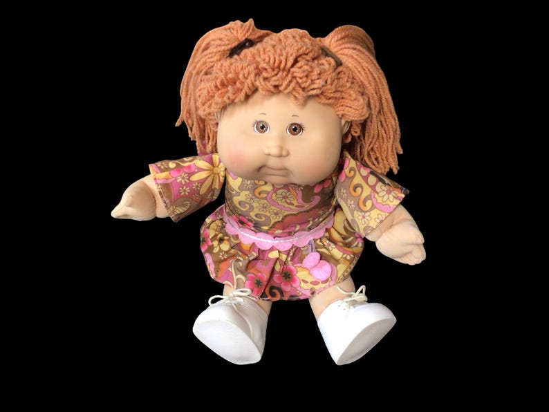 Puede incluir: Mu&ntilde;eca vintage con cabello de hilo naranja peinado en coletas. La mu&ntilde;eca viste un vestido con estampado floral en tonos rosas y marrones, y zapatos blancos. La mu&ntilde;eca tiene una cara redonda y est&aacute; sobre un fondo negro.