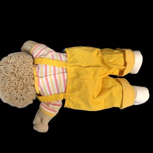 K&ouml;nnte beinhalten: Eine Vintage-Puppe mit hellbrauner, lockiger Frisur. Die Puppe tr&auml;gt ein gestreiftes Hemd mit rosa und gelben Streifen, eine gelbe Latzhose und wei&szlig;e Schuhe. Die Puppe liegt auf schwarzem Hintergrund.