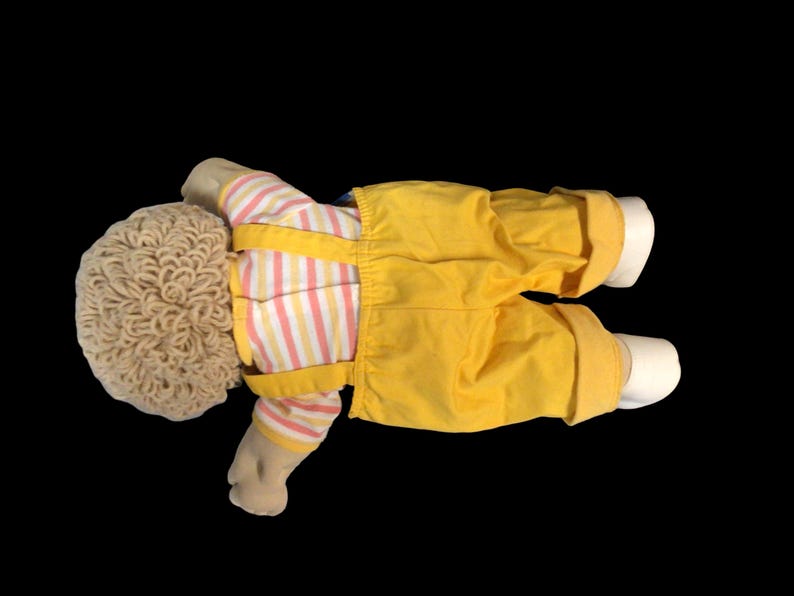 K&ouml;nnte beinhalten: Eine Vintage-Puppe mit hellbrauner, lockiger Frisur. Die Puppe tr&auml;gt ein gestreiftes Hemd mit rosa und gelben Streifen, eine gelbe Latzhose und wei&szlig;e Schuhe. Die Puppe liegt auf schwarzem Hintergrund.