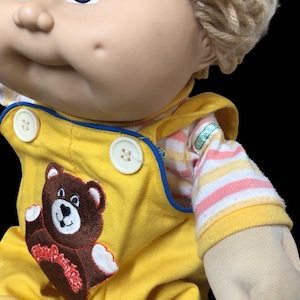 K&ouml;nnte beinhalten: Eine Vintage-Puppe mit blonden Garnhaaren und blauen Augen. Die Puppe tr&auml;gt einen gelben Overall mit einem braunen Teddyb&auml;r-Applikation und einem gestreiften Hemd. Der Overall hat wei&szlig;e Kn&ouml;pfe und blaue Paspeln.