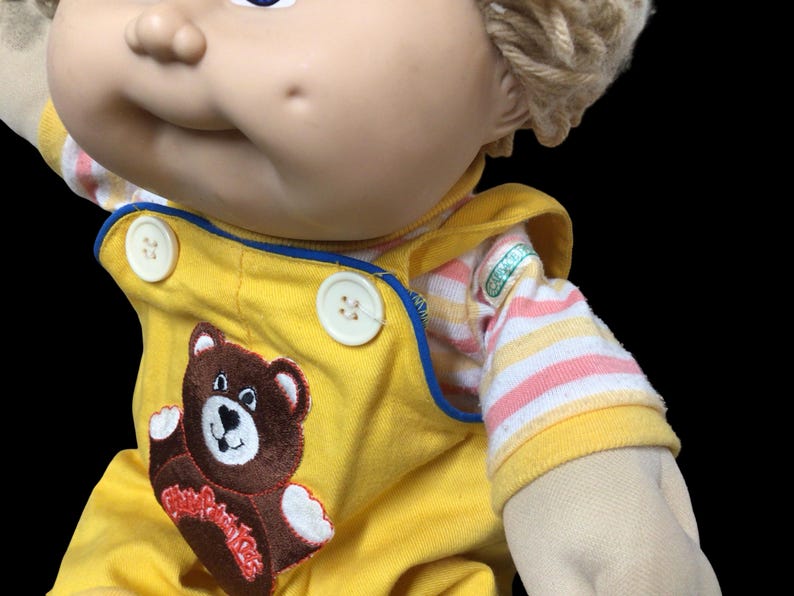 K&ouml;nnte beinhalten: Eine Vintage-Puppe mit blonden Garnhaaren und blauen Augen. Die Puppe tr&auml;gt einen gelben Overall mit einem braunen Teddyb&auml;r-Applikation und einem gestreiften Hemd. Der Overall hat wei&szlig;e Kn&ouml;pfe und blaue Paspeln.
