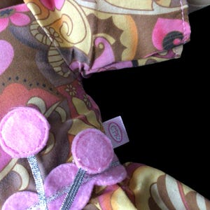 Puede incluir: Primer plano de una tela con un estampado floral retro en tonos rosa, marr&oacute;n y amarillo. Se ven adornos de flores de fieltro rosa con detalles de cinta plateada. Tambi&eacute;n es visible una etiqueta con las letras "CPK".