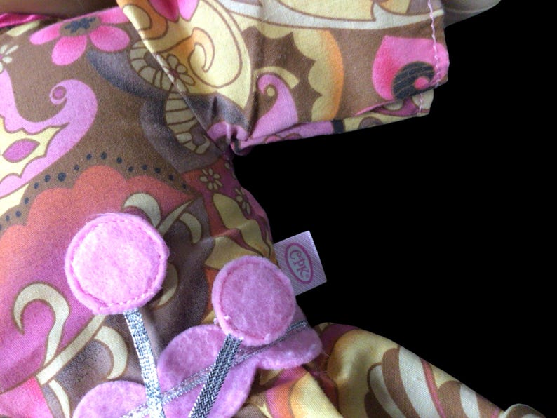 Puede incluir: Primer plano de una tela con un estampado floral retro en tonos rosa, marr&oacute;n y amarillo. Se ven adornos de flores de fieltro rosa con detalles de cinta plateada. Tambi&eacute;n es visible una etiqueta con las letras "CPK".