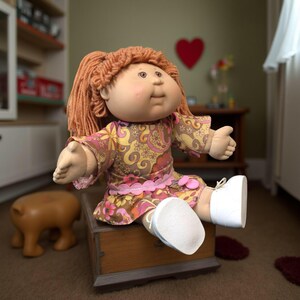 Puede incluir: Una mu&ntilde;eca Cabbage Patch Kid vintage con cabello de hilo rojo, vestida con un vestido floral en tonos de amarillo, rosa y marr&oacute;n. La mu&ntilde;eca est&aacute; sentada en un cofre de madera y lleva zapatos blancos. Los brazos de la mu&ntilde;eca est&aacute;n extendidos.