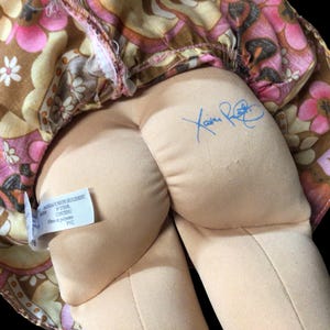 Puede incluir: Una mu&ntilde;eca vintage con un vestido estampado floral en tonos rosa, marr&oacute;n y amarillo. El cuerpo de la mu&ntilde;eca es beige y se ve una firma azul. Tambi&eacute;n hay una etiqueta con texto.