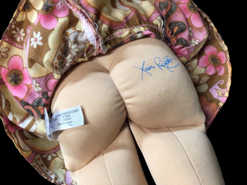 Puede incluir: Una mu&ntilde;eca vintage con un vestido estampado floral en tonos rosa, marr&oacute;n y amarillo. El cuerpo de la mu&ntilde;eca es beige y se ve una firma azul. Tambi&eacute;n hay una etiqueta con texto.