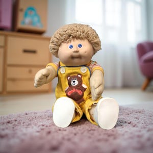 K&ouml;nnte beinhalten: Eine Cabbage Patch Kids-Puppe mit blondem, lockigem Haar und blauen Augen. Die Puppe tr&auml;gt einen gelben Overall mit einem braunen B&auml;renaufn&auml;her und wei&szlig;en Schuhen. Die Puppe sitzt auf einem rosa Teppich.